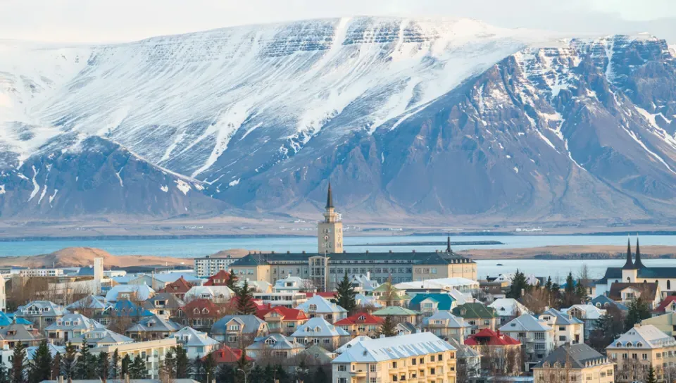 A town with colorful buildings is set before a snowy mountain range under a cloudy sky